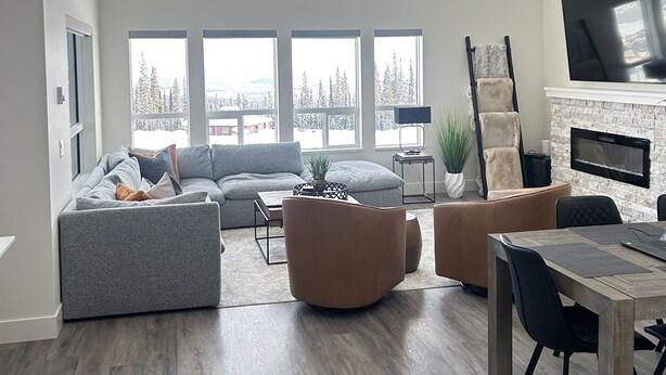 Un salon spacieux avec un grand canapé d’angle, une table basse et un coin repas comprenant une table et des chaises en bois. De nombreuses fenêtres laissent entrer la lumière naturelle et offrent une vue sur les arbres.