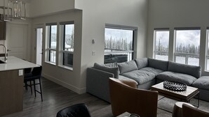 Un vaste espace de vie comprenant un canapé d’angle gris, une table basse en bois et une vue sur un paysage enneigé, visible à travers de grandes fenêtres.
