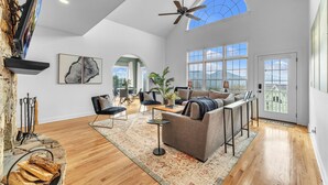 A spacious living room with a high ceiling, a ceiling fan, a large window, and a fireplace. - 7br Asheville Mansion, Views, Pool, Full Spa, Hot Tub, Gameroom - close to DownT (Asheville)