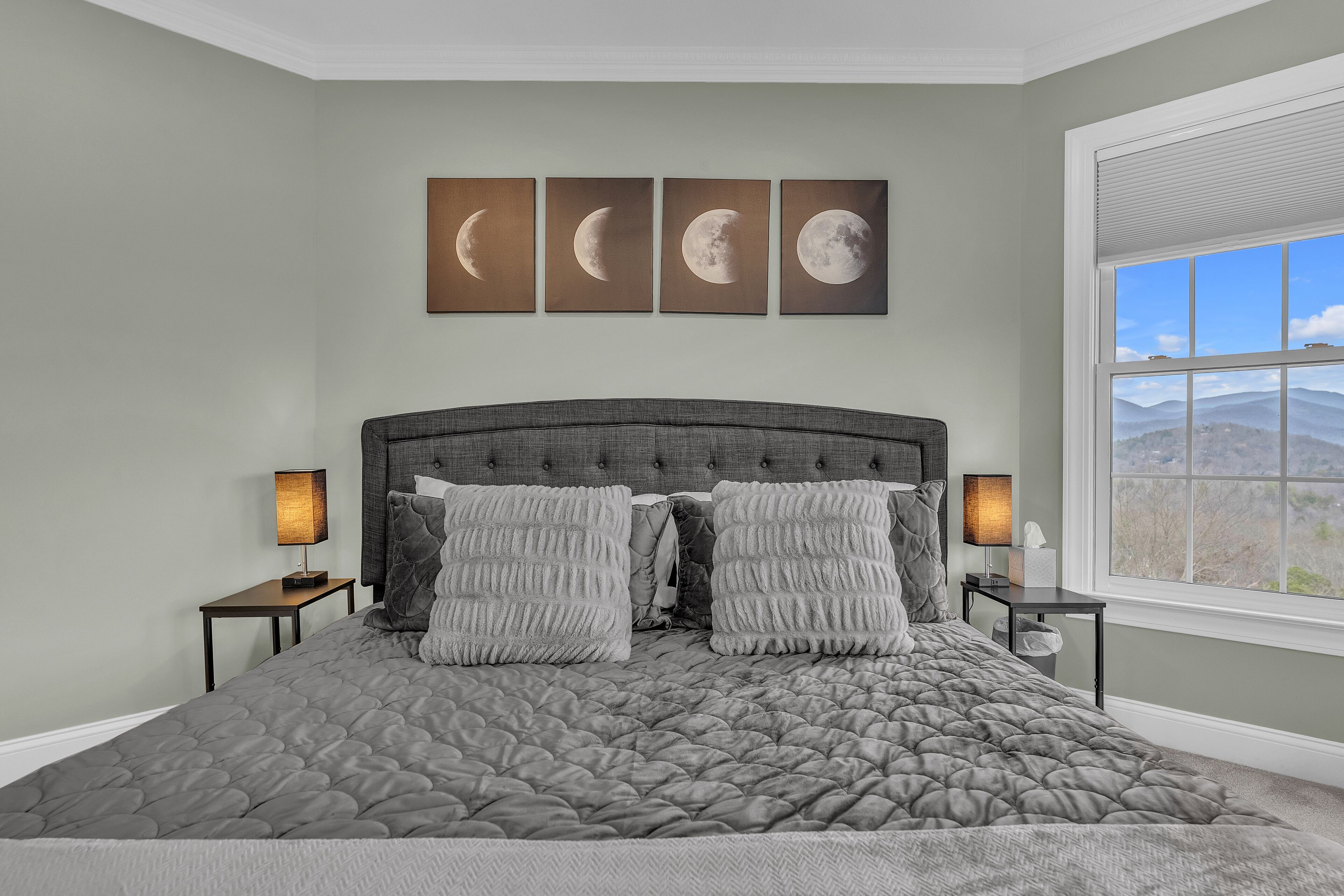Un dormitorio con una cama grande, dos mesitas de noche, un ventanal con vistas a la montaña y cuatro cuadros con las fases de la luna en la pared.