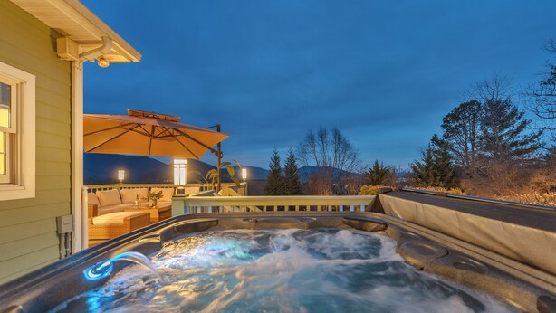 A hot tub with a waterfall feature, surrounded by a deck with outdoor furniture and a large umbrella. - 7br Asheville Mansion, Views, Pool, Full Spa, Hot Tub, Gameroom - close to DownT (Asheville)