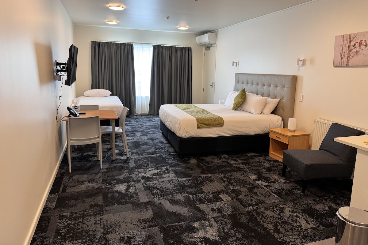 A hotel room with a bed, a desk with a chair, a small table, and a radiator.