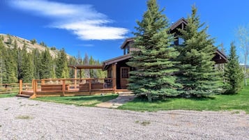 A lodge with a wooden deck, a large evergreen tree, and a mountainous backdrop.