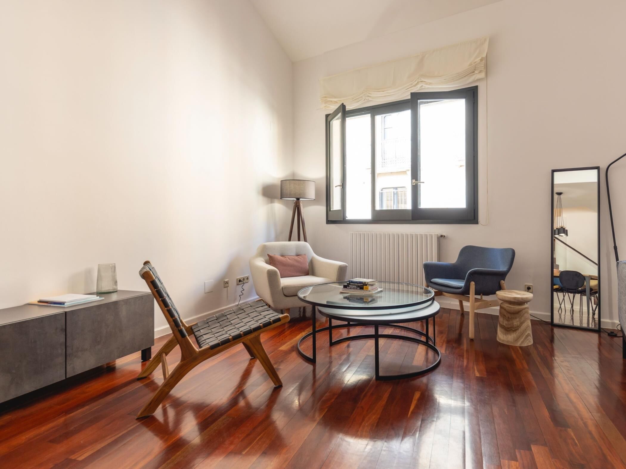 A modern living room with a wooden floor, a round coffee table, a sofa, a chair, a small side table, a lamp, and a window with curtains.