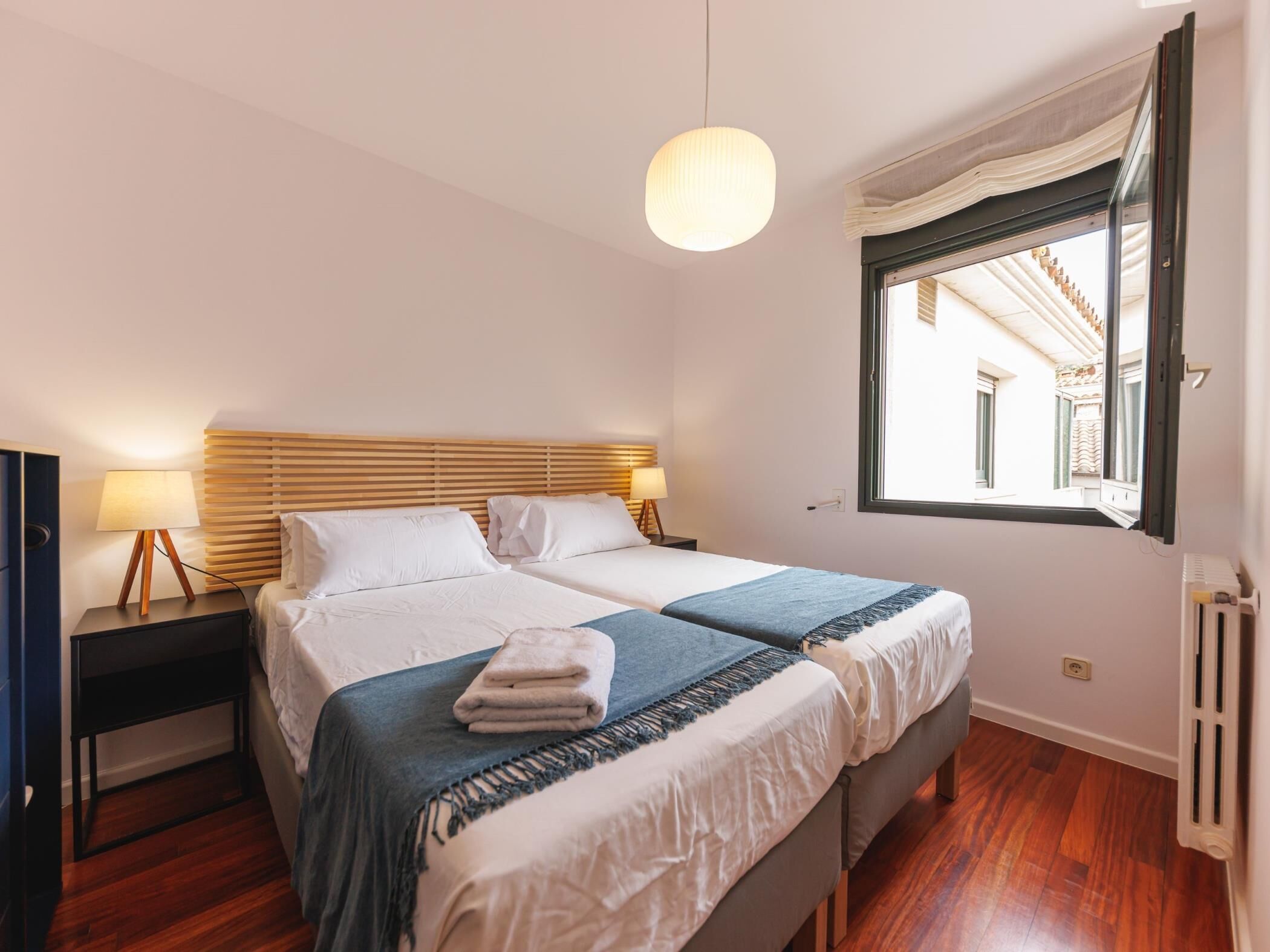 A hotel room with two beds, a wooden headboard, a bedside table with a lamp, a window with a view of buildings, and a radiator.