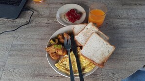 Une assiette de petit-déjeuner avec des toasts, des œufs brouillés et des légumes, un verre de jus d’orange et un petit bol de confiture, le tout posé sur une table en bois.