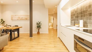 A modern kitchen with a built-in oven, a dining area with a wooden table and chairs, and a hallway leading to a bathroom.