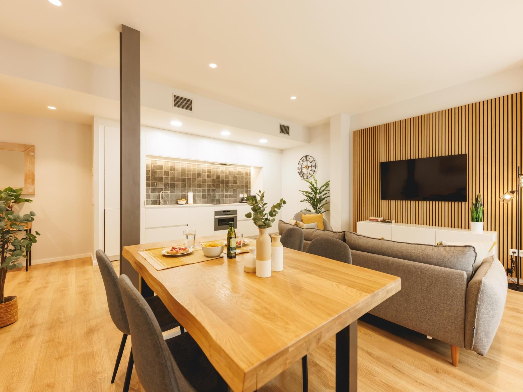 Ein modernes Wohnzimmer mit einem Holzesstisch, einem grauen Sofa und einem an einer Holzpaneelwand montierten Fernseher.
