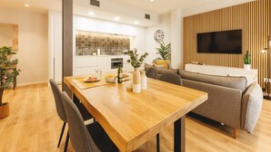 A modern living room with a wooden dining table, grey sofa, and a TV mounted on a wooden panel wall.