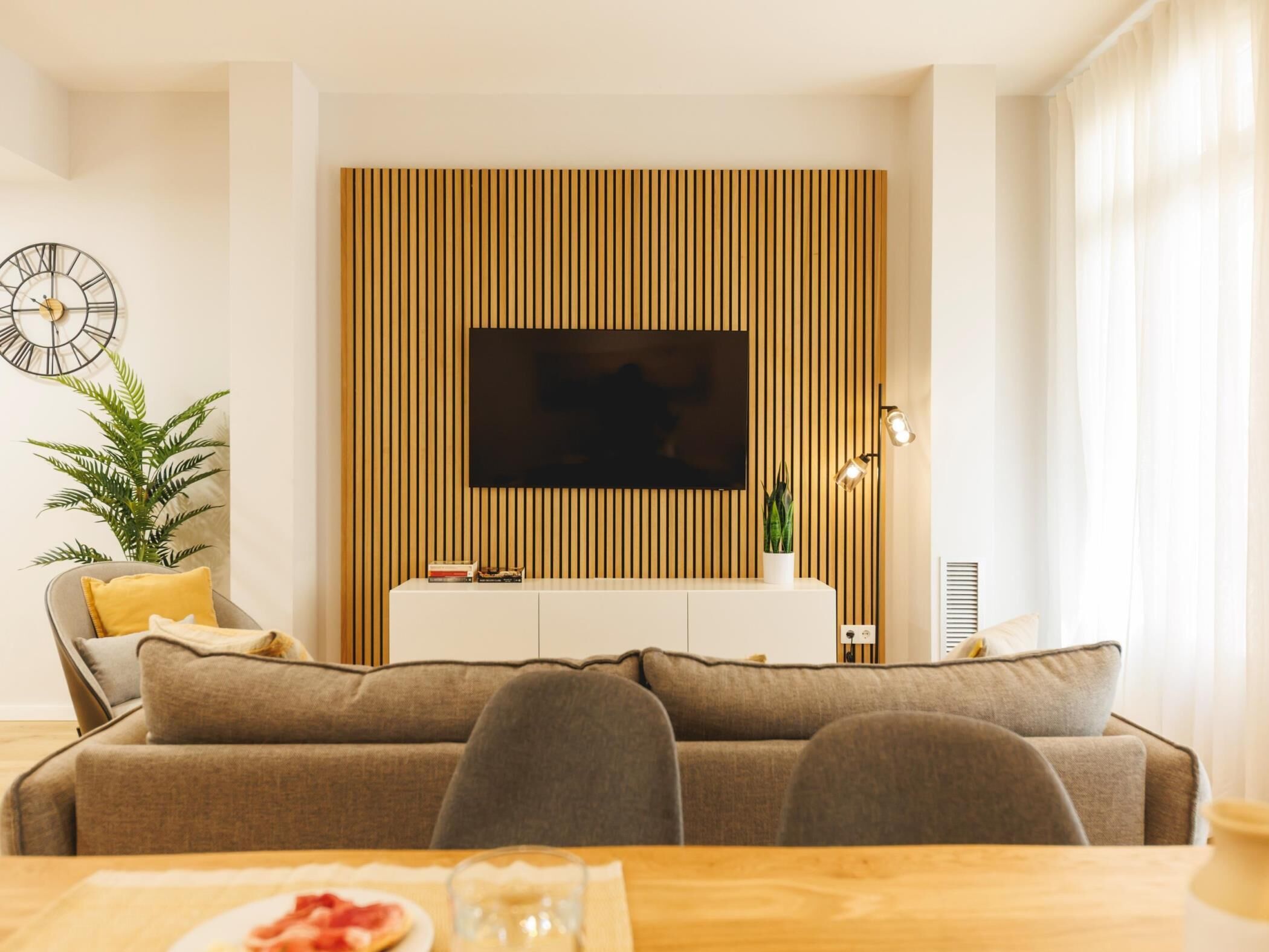 Ett modernt vardagsrum med en stor platt-TV som är monterad på en vägg med träribbor, en soffa, ett matbord med en tallrik mat på och en krukväxt.