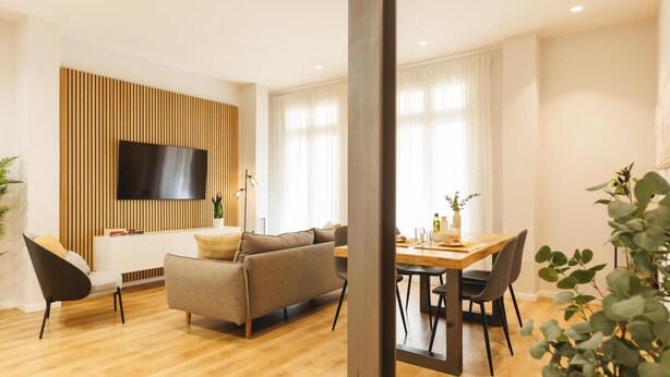 A modern living room with a sofa, a dining table, and a TV mounted on a wooden panel wall.