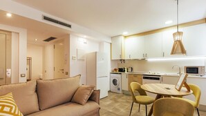Una cocina moderna con armarios blancos, un comedor con una mesa redonda y sillas amarillas, y un sofá beige con cojines estampados.