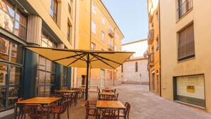 Un café al aire libre con mesas y sillas de madera, un paraguas grande y edificios con balcones y ventanas al fondo.