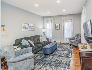A modern living room with a grey sofa, a blue ottoman, and a grey armchair. There is a large flat-screen TV on a stand, a lamp, and a framed picture on the wall.