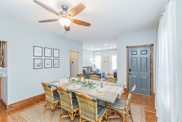 A dining area with a table set for six, surrounded by wicker chairs, a ceiling fan, and framed artwork on the walls.