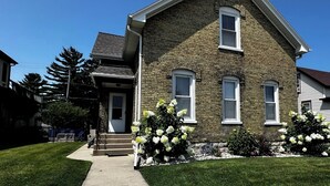 A two-story brick house with a front porch, a visible front door, and a small front yard with white flowering plants. - Family Retreat Near Lake Michigan | Extended Work Stay (Racine)
