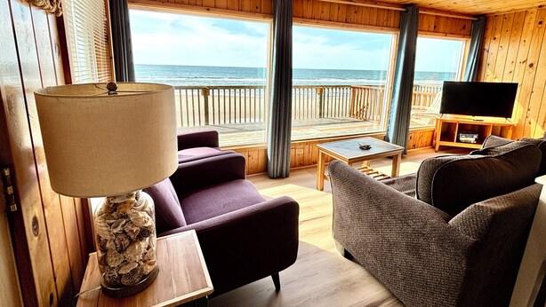 Et interiør af en træhytte med et stort vindue der kigger ud over havet, møbleret med en sofa, lænestol og et tv-skab.