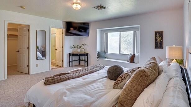 Quarto com uma cama grande, televisão na parede, janela e mesa de cabeceira com luminária.