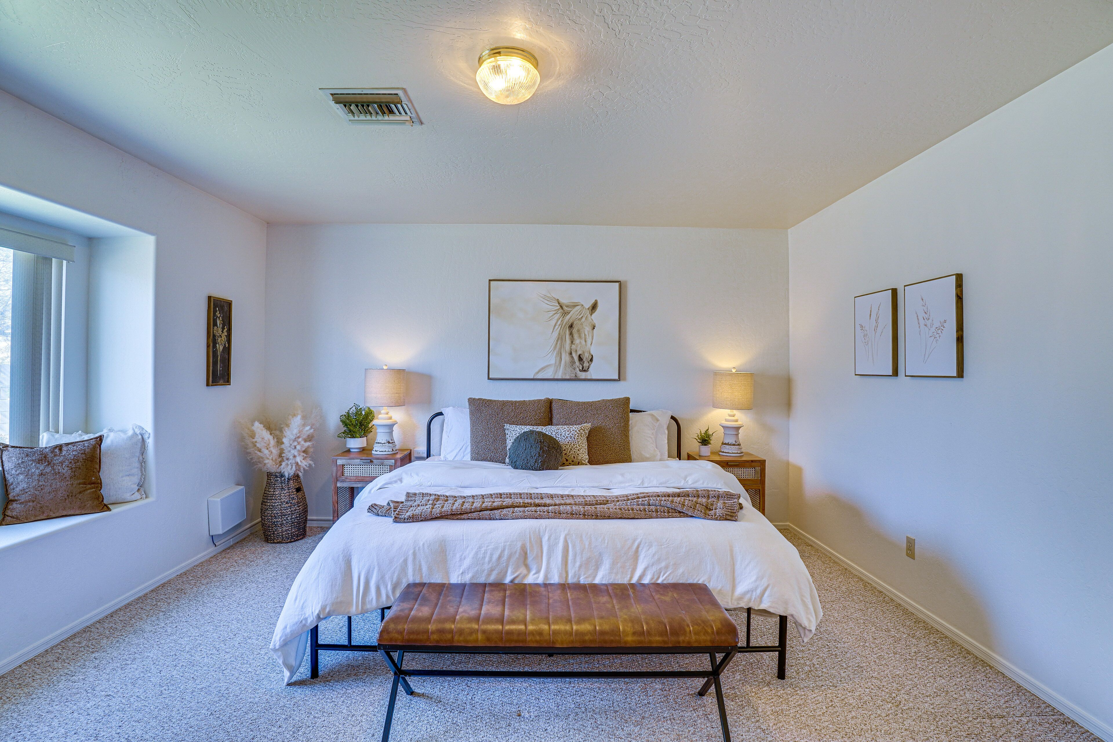 A bedroom with a large bed, bedside lamps, a window with blinds, a bedside plant, and a wall-mounted artwork.