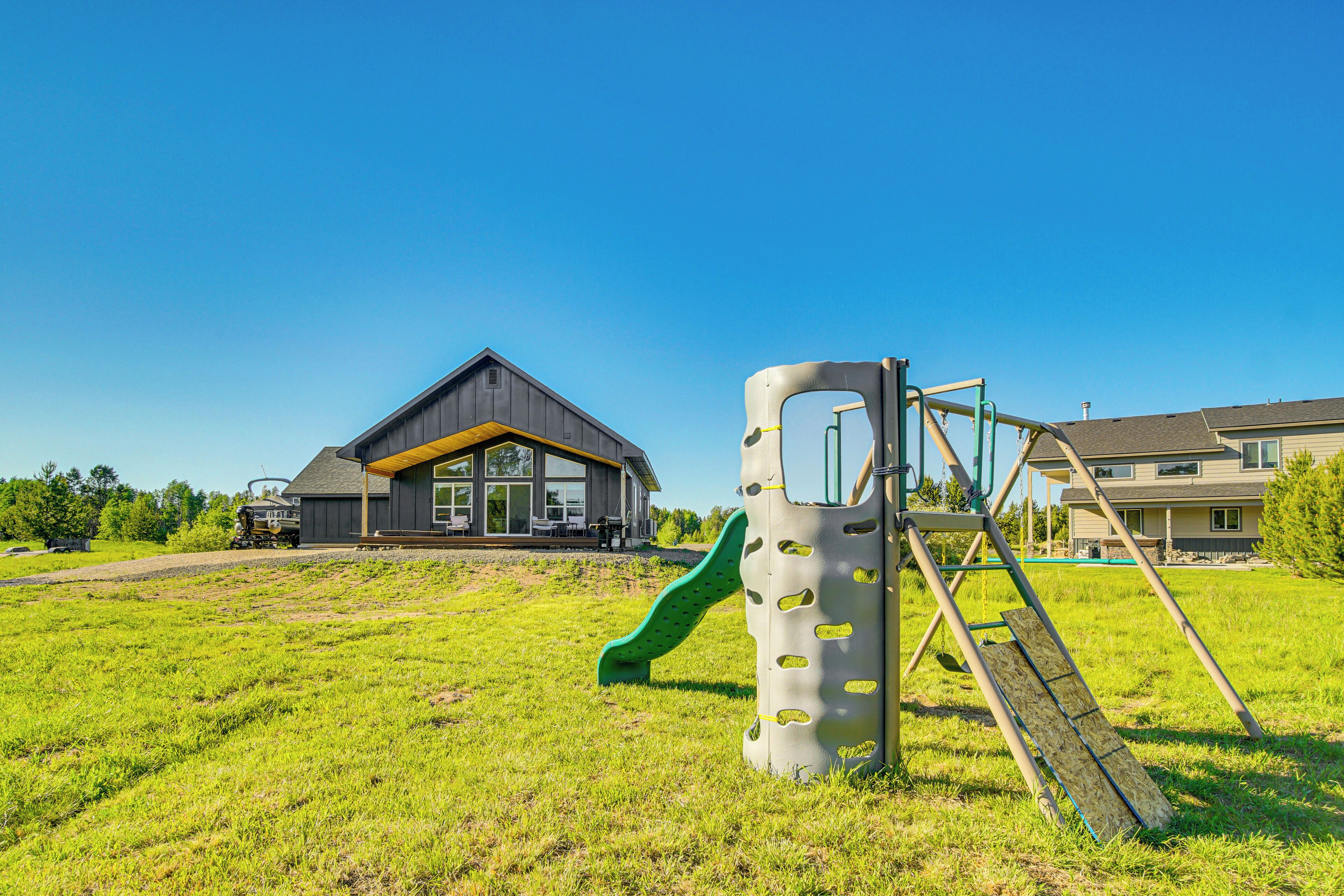 A playground with a slide and climbing structure, a house with a dark roof and yellow trim, and a clear blue sky.