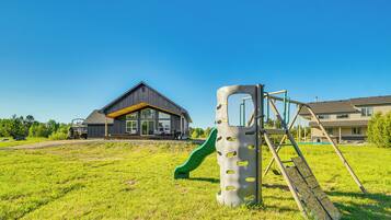 A playground with a slide and climbing structure, a house with a dark roof and yellow trim, and a clear blue sky.
