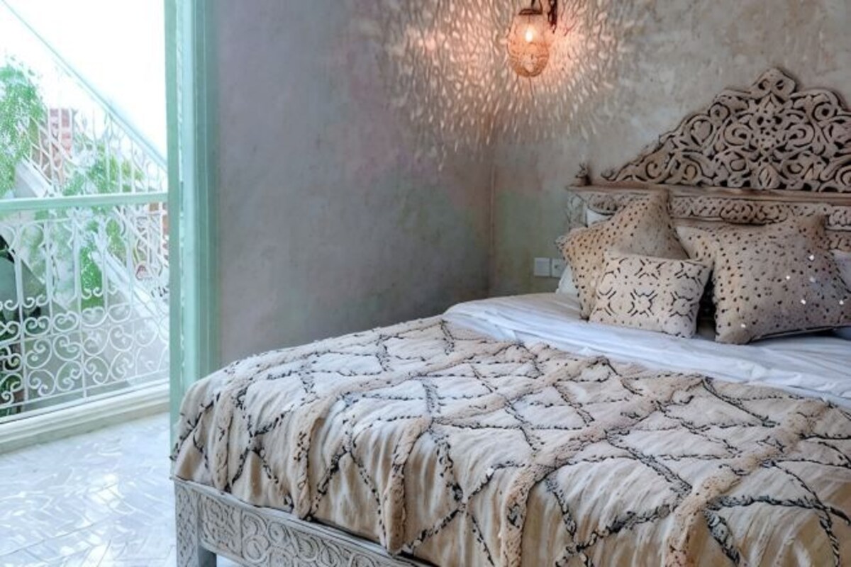 A bedroom with a bed, patterned bedding, a decorative headboard, and a balcony with a railing.