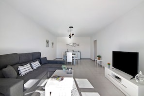 Un salon moderne avec un canapé gris, un téléviseur à écran plat et un coin repas à l’arrière-plan.