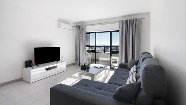 En moderne stue med et fladskærms-tv, en grå sofa og en balkon med gardiner.