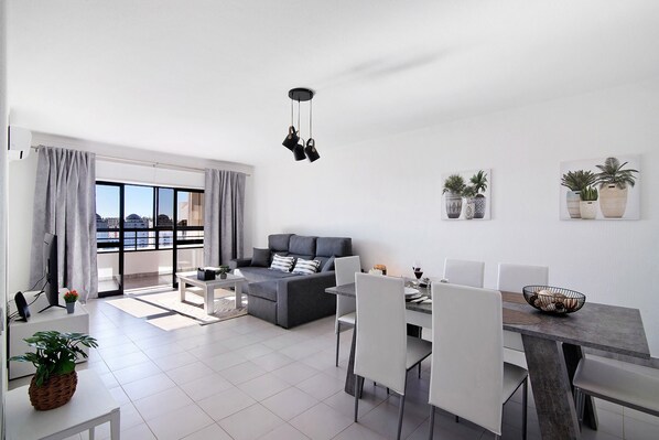 Een moderne woonkamer met een grijze bank, een eettafel en stoelen, een balkon met gordijnen en potplanten aan de muren.