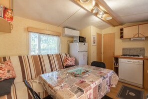 A compact kitchenette with a microwave, refrigerator, and dishwasher. There is a dining table with a floral tablecloth and a bench with striped cushions.
