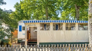 A mobile home with a blue and white striped awning, a covered porch, and a fenced yard. - Mobile Home 'Mh 100 La Tremblade' with Shared Pool, Terrace, and Private Garden (La Tremblade)