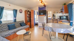 A compact RV interior with a sofa, a small dining table, a kitchenette, and a TV. - Mobile Home 'Mh 100 La Tremblade' with Shared Pool, Terrace, and Private Garden (La Tremblade)