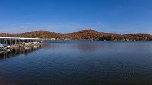 A calm lake with docked boats, a covered pier, and a forested hillside in the background. - Amazing view from water-front condo! Close to golf courses and great dining. (Camdenton)