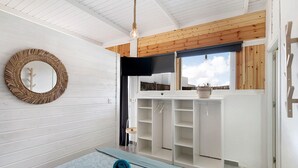 Una habitación con techo de madera, un espejo circular tejido, una cama con colcha azul estampada y un armario con las puertas abiertas.
