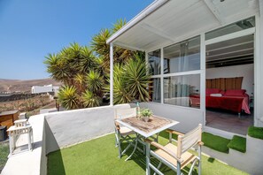 Um pátio moderno ao ar livre com mesa e cadeiras, uma cama vermelha visível por uma porta de vidro e vista para a paisagem.