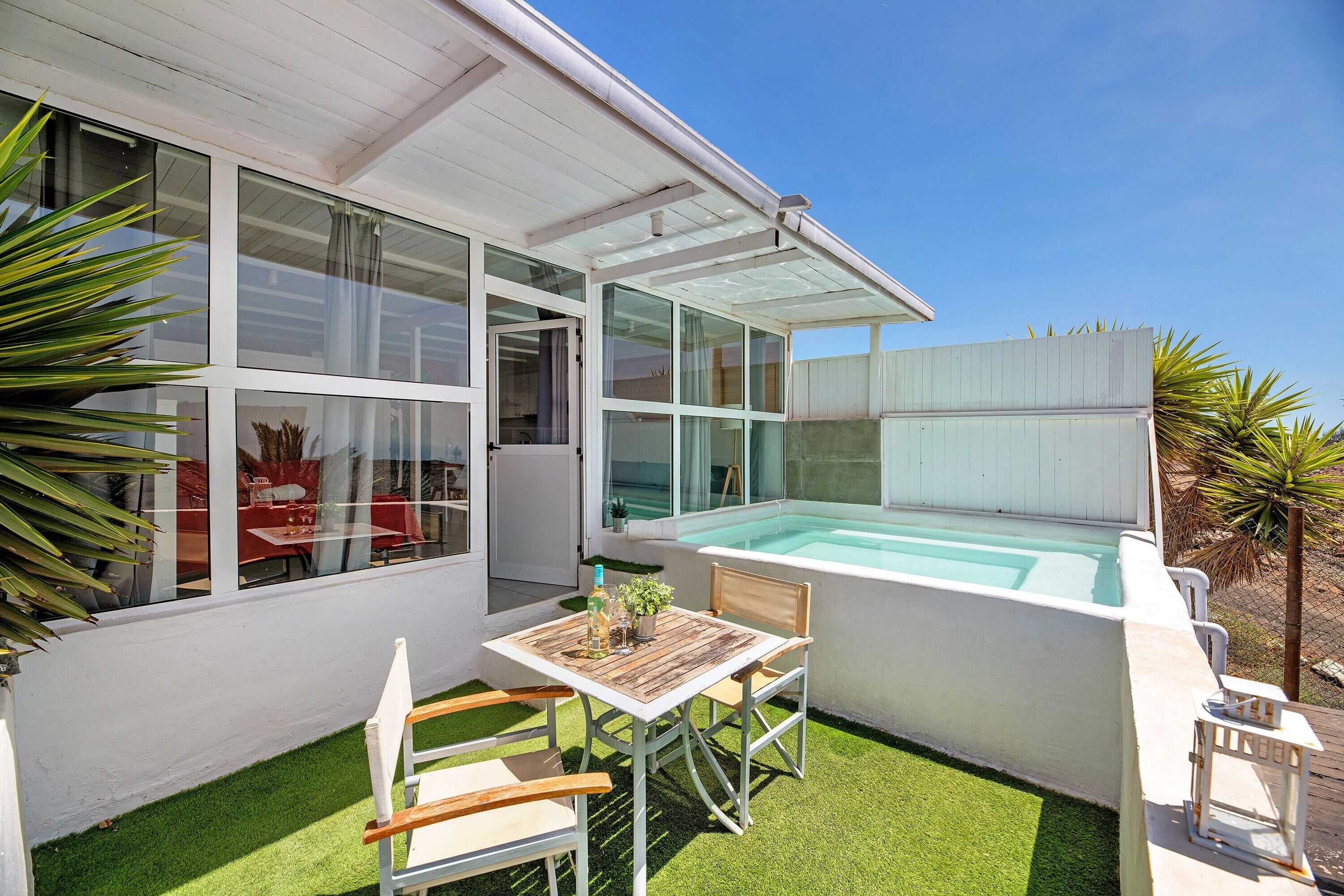Een modern huis met een glazen hottub, een klein terras met een tafel en stoelen, en een verzorgd gazon.