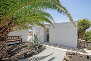 Um edifício branco com um caminho de pedra, um banco e diversas plantas.