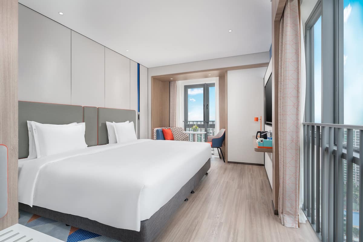 A modern hotel room with a large bed, a desk with a chair, a balcony with a railing, and a large window offering a view of the city.