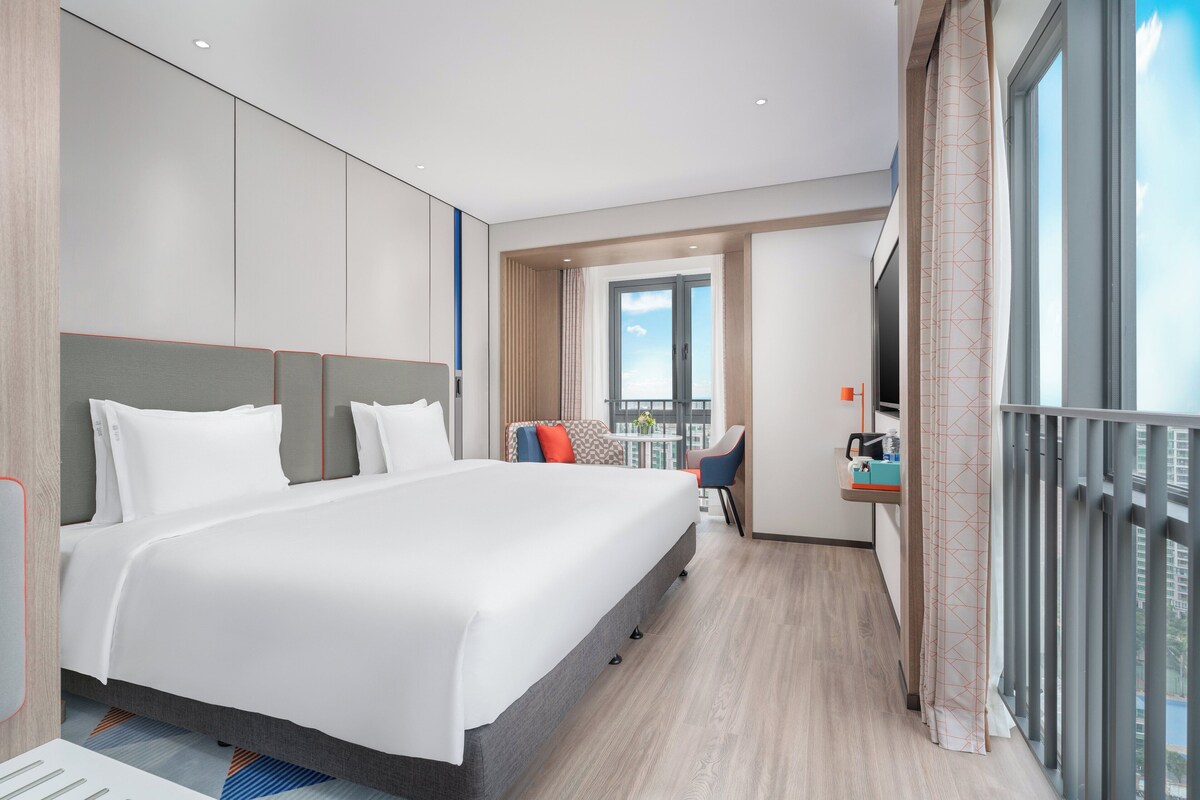 A modern hotel room with a large bed, a desk with a chair, a balcony with a railing, and a large window offering a view of the city.