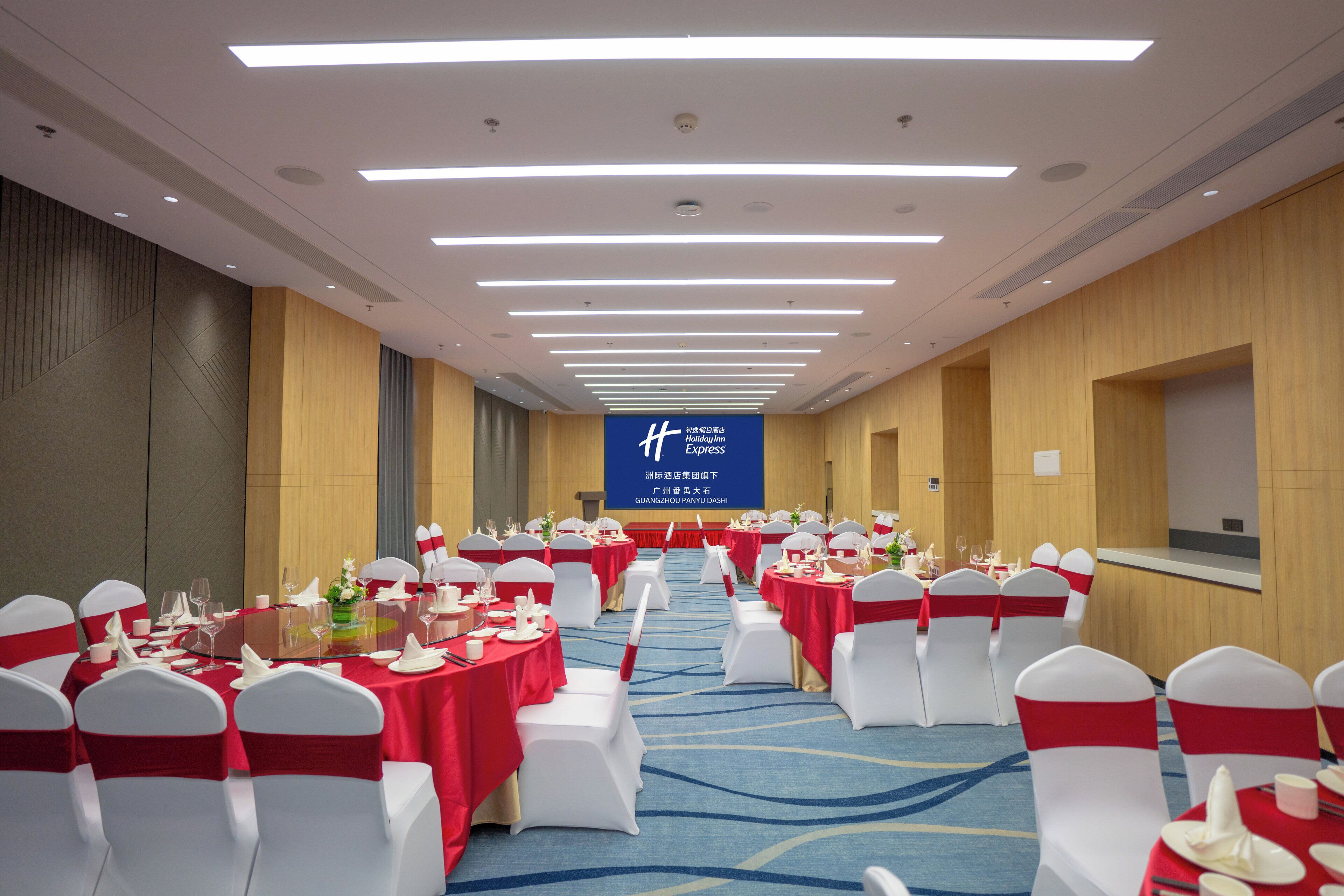 Un salón de banquetes con filas de mesas redondas, sillas blancas y lazos rojos. Una pantalla grande muestra texto en un idioma extranjero.