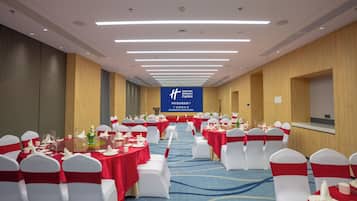 Un salón de banquetes con filas de mesas redondas, sillas blancas y lazos rojos. Una pantalla grande muestra texto en un idioma extranjero.
