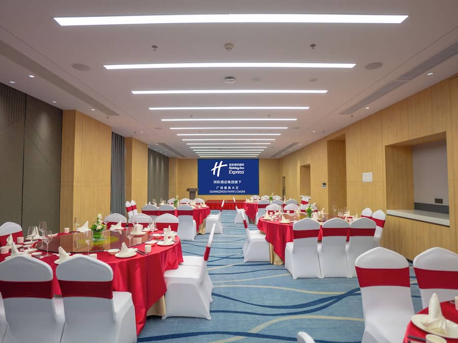 Un salón de banquetes con filas de mesas redondas, sillas blancas y lazos rojos. Una pantalla grande muestra texto en un idioma extranjero.