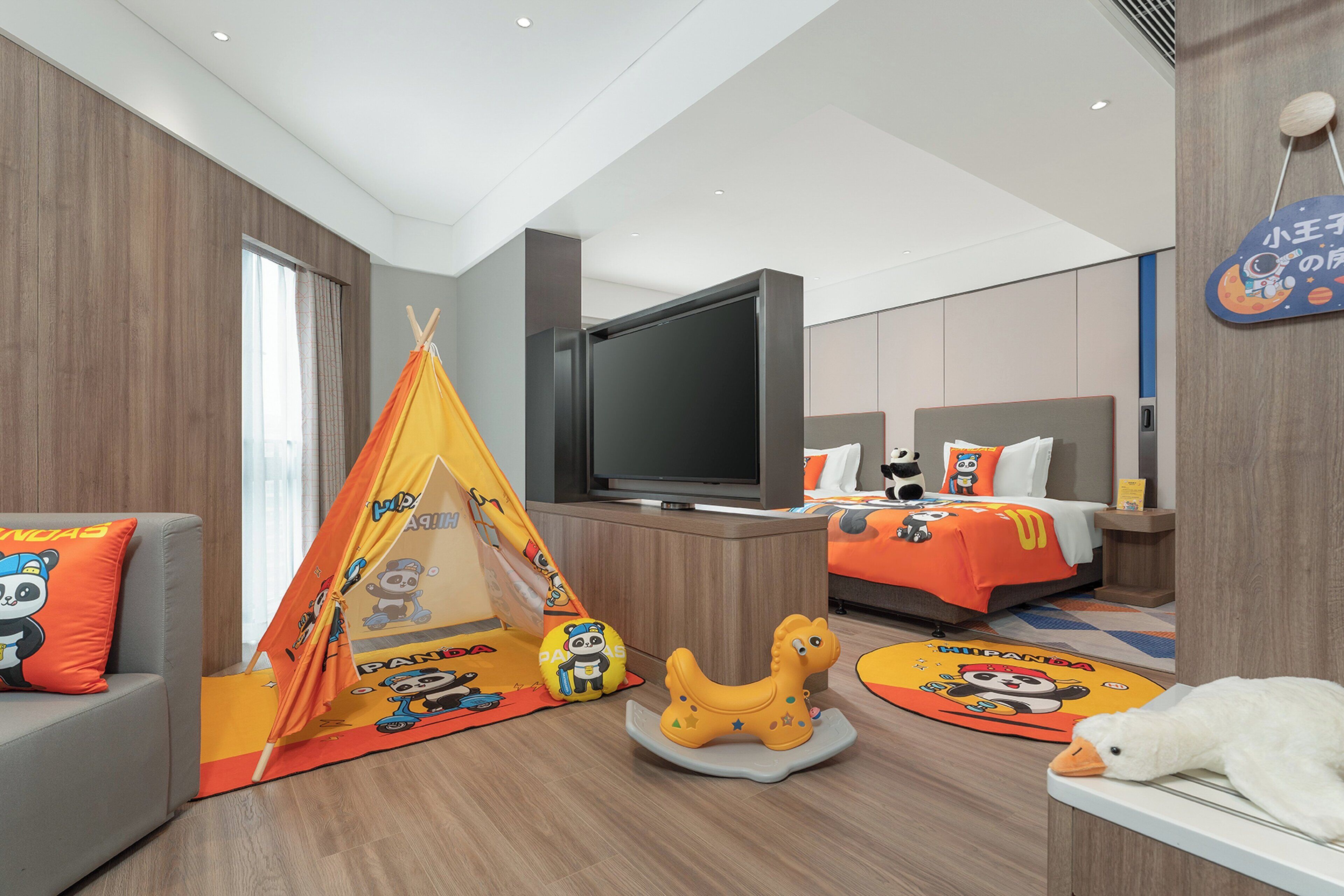 Quarto de hotel moderno com uma cama grande, televisão, sofá, tenda infantil e um balanço de cavalo em forma de pato.