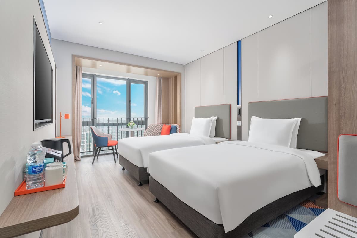 A hotel room with two beds, a desk with a coffee maker, a TV, and a balcony with a view of the sky.