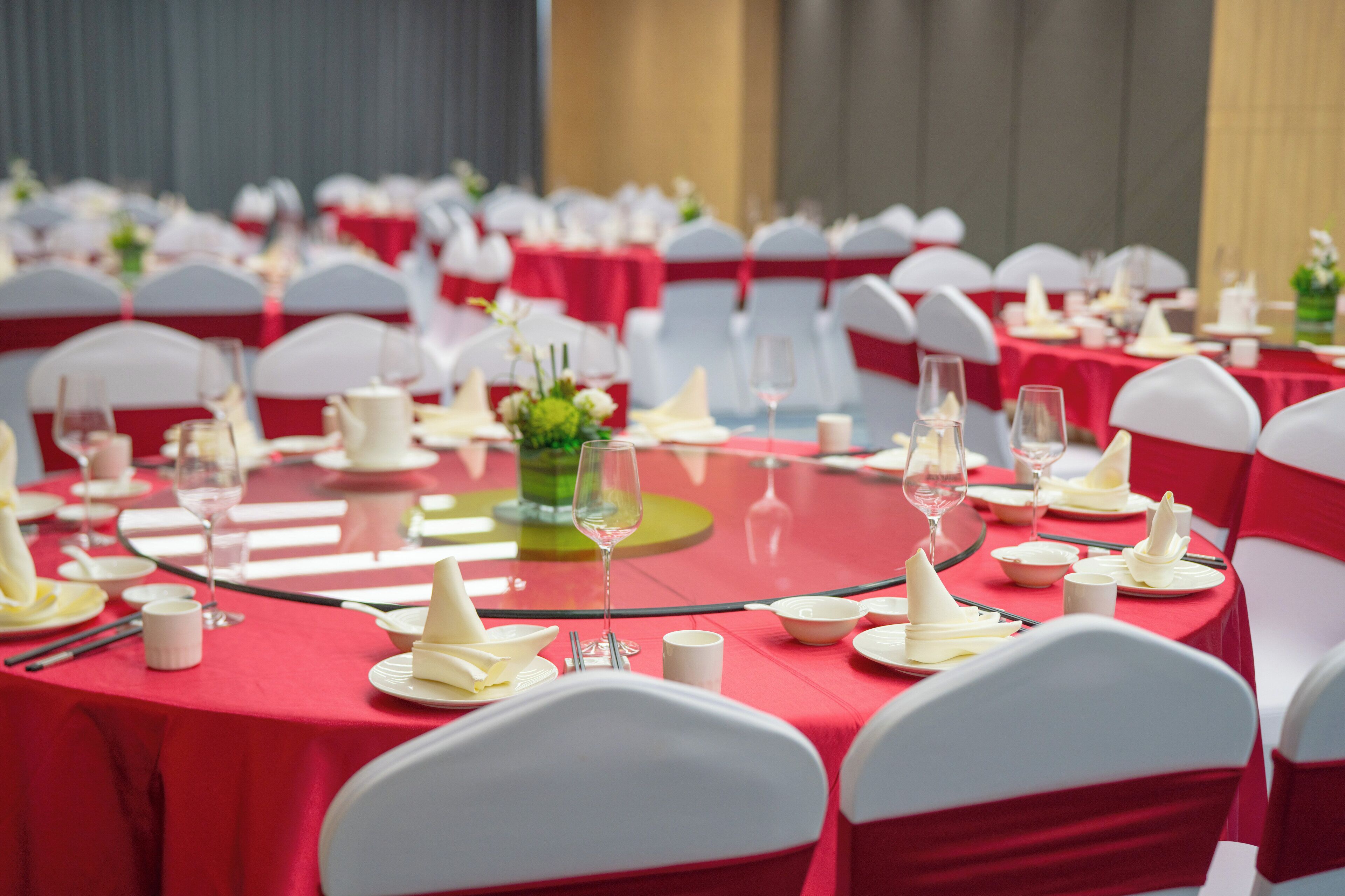 A banquet hall with round tables set for a formal event, featuring red tablecloths and white chairs with red sashes.