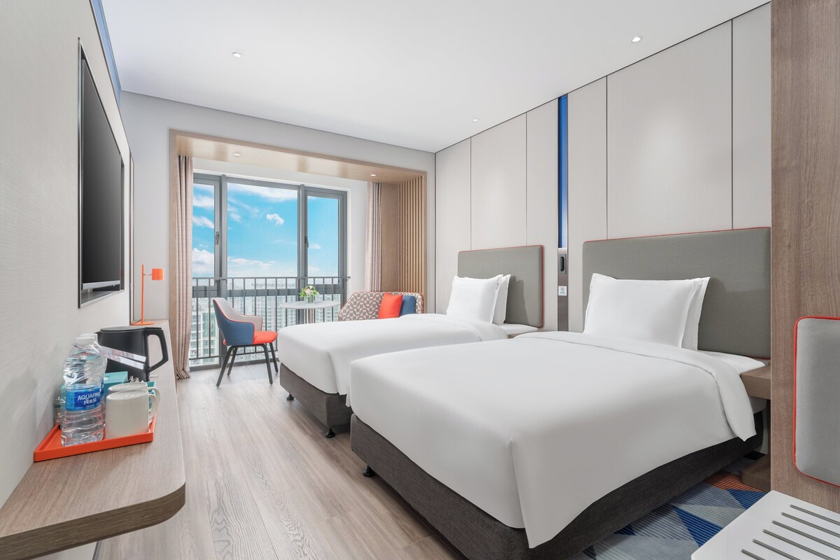 A hotel room with two beds, a desk with a coffee maker, a TV, and a balcony with a view of the sky.