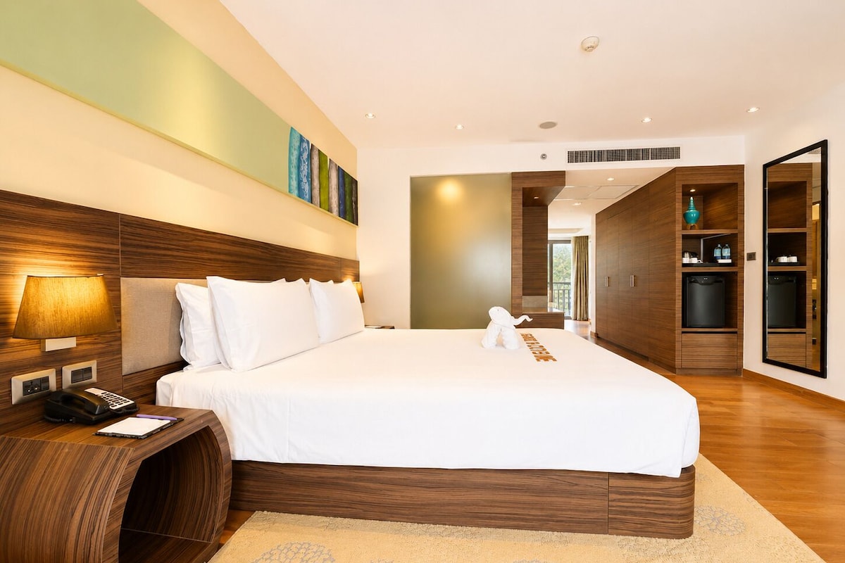 Quarto de hotel moderno com uma cama grande, cabeceira de madeira, uma escrivaninha com luminária e um minibar.