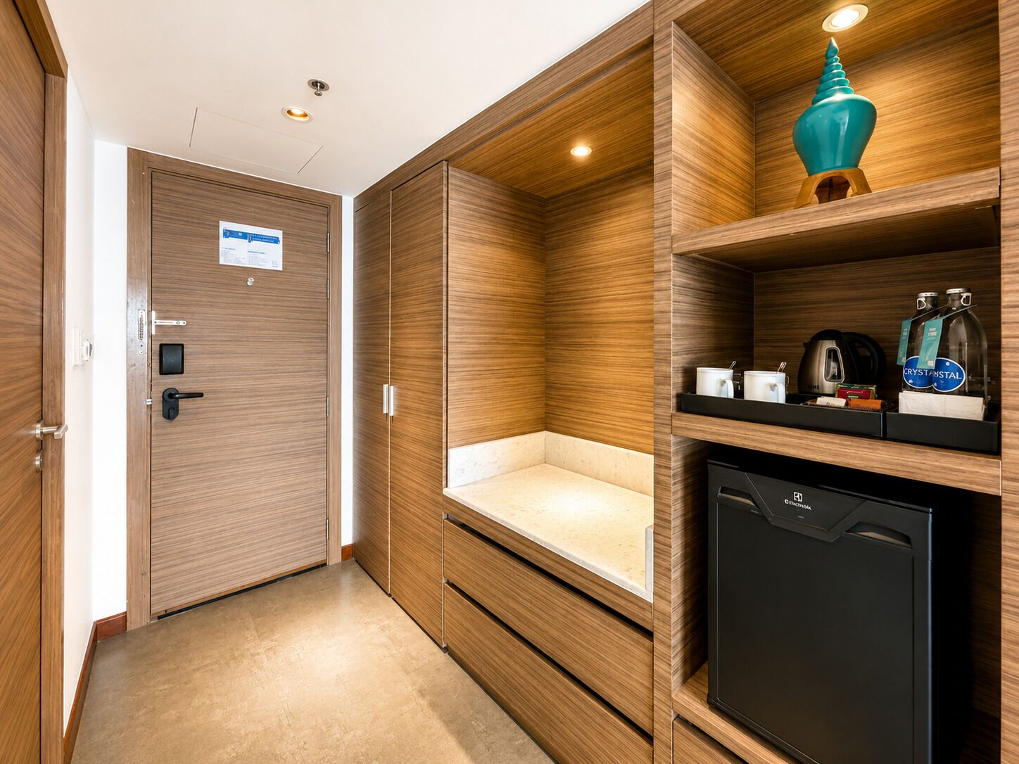 A hotel room with a wooden wardrobe, a minibar, a small refrigerator, and a shelf with toiletries.