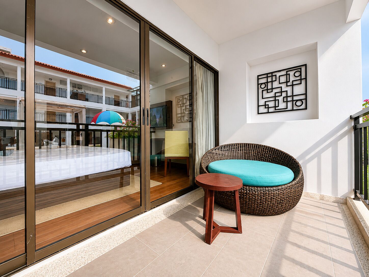A balcony with a wicker sofa, a small wooden stool, and a glass door leading to a balcony with a view of a building.