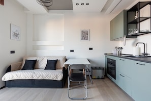 Un espace de vie compact comprenant un lit, une table à manger, une chaise, un coin cuisine avec des placards et un évier, ainsi que des œuvres d’art accrochées aux murs.